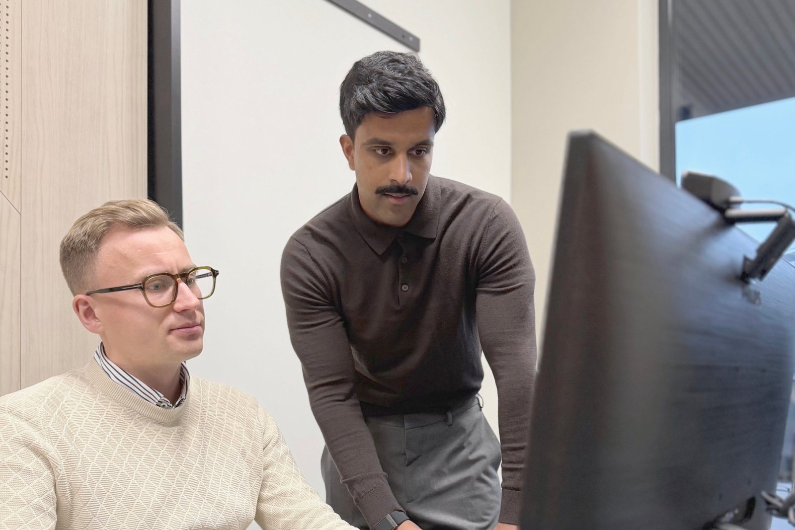 Inthujan Rasalingam leaning over a desk as Kjell Petersen Synstad works on a large monitor in the Capacity Connect office