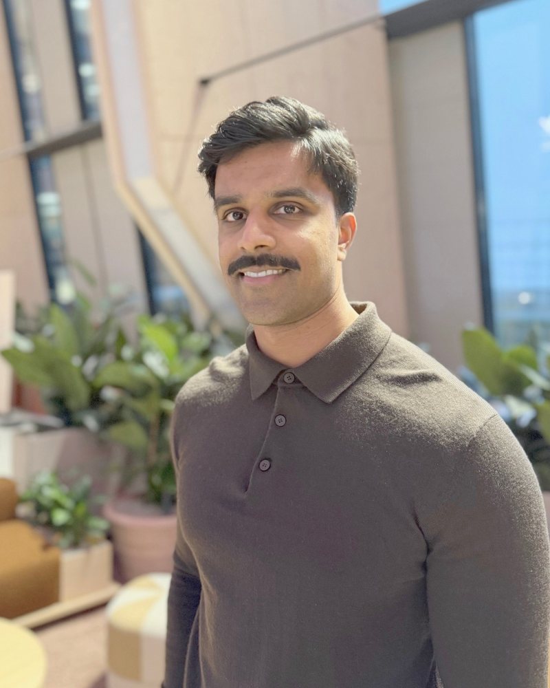Portrait of Inthujan Rasalingam smiling, with plants and tall windows behind him